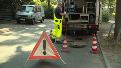 Ein Mitarbeiter der Berliner Wasserbetriebe arbeitet auf einer Straße an einem Gulli. Er steht vor einem Fahrzeug. Im Vordergrund ist ein Warnschild zu sehen.