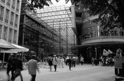 Stahl-Glas Architektur des Arkaden im Hintergrund, davor viele Passanten, links Cafebetrieb, rechts eine Gruppe von Menschen