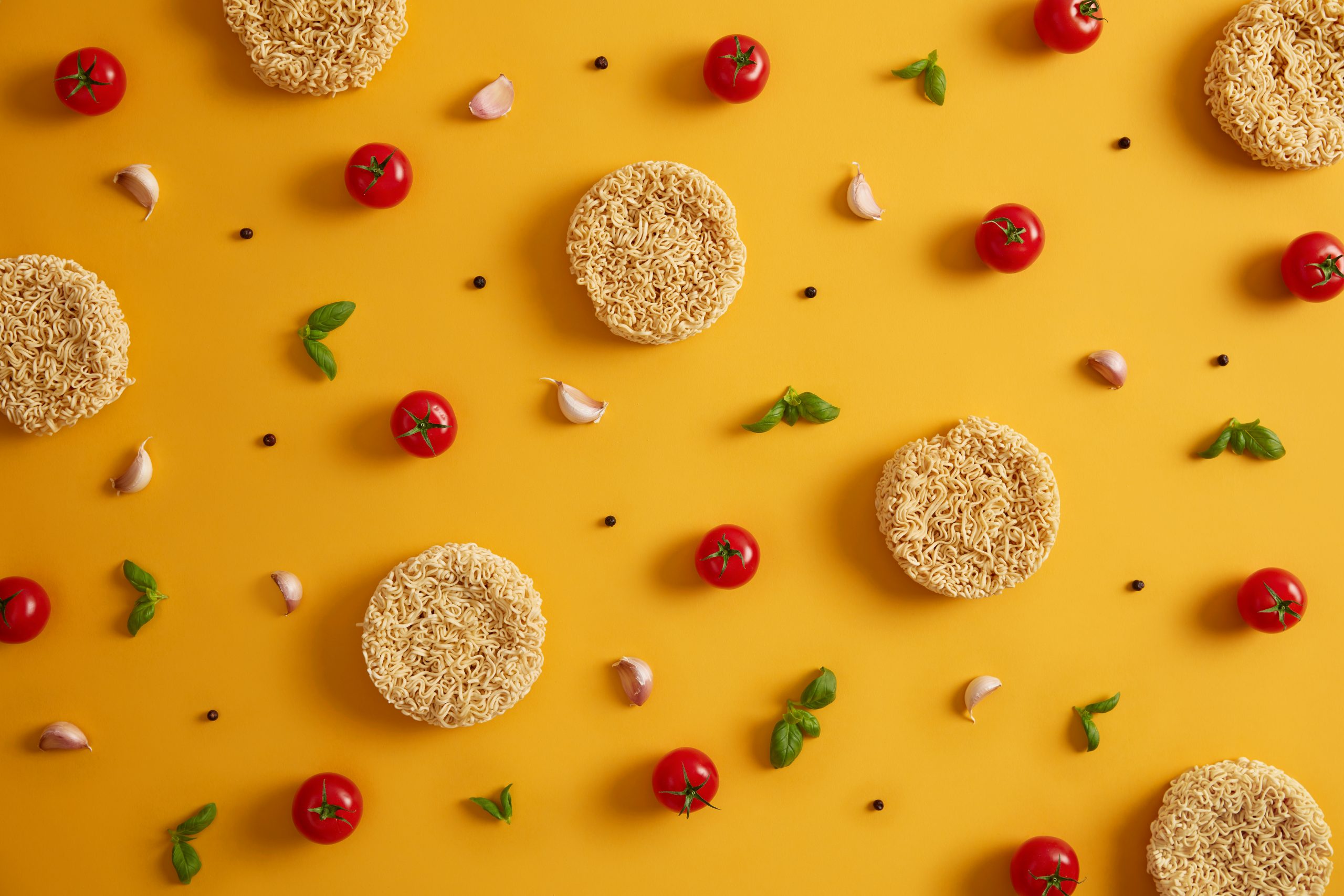 Dry instant noodles with tomatoes, garlic, basil and pepper for preparing fresh soup, yellow background. Preparing qucik lunch. Unhealthy eating and fast food concept. Ingredients for making dish